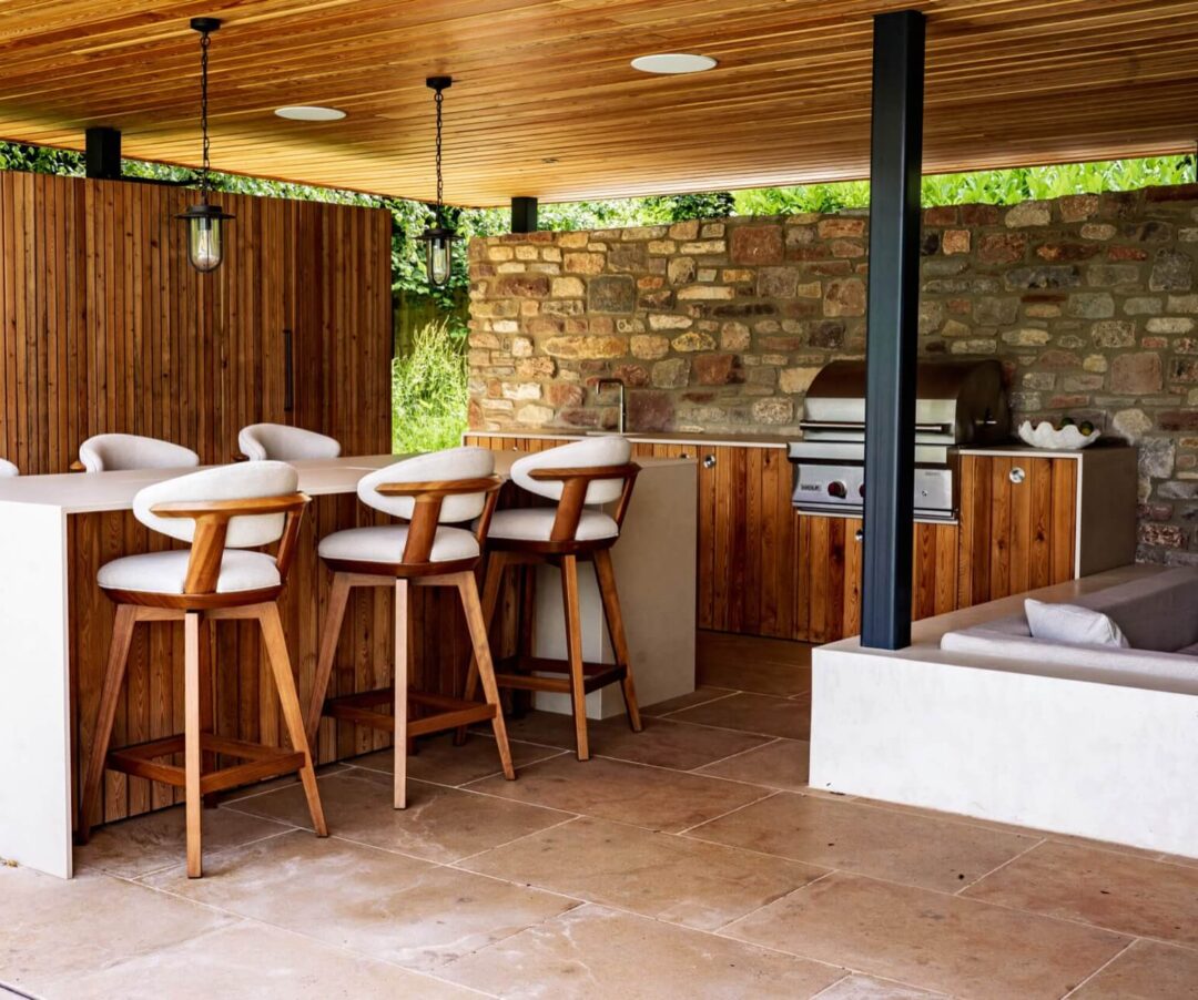 Bespoke wood-panelled pool house by Artisan Landscapes, with kitchen island seating, outdoor kitchen and grill area.