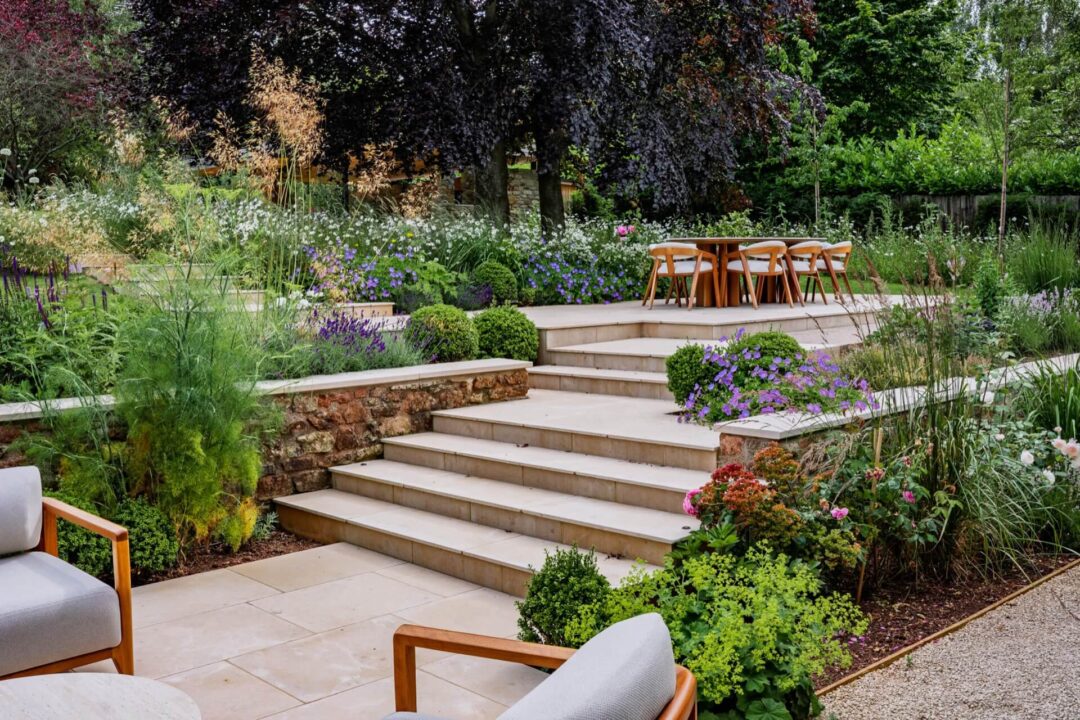 Looking from a paved seating area up the steps to further terraces, with dining area, and colourful natural planting, and copper beech trees beyond.