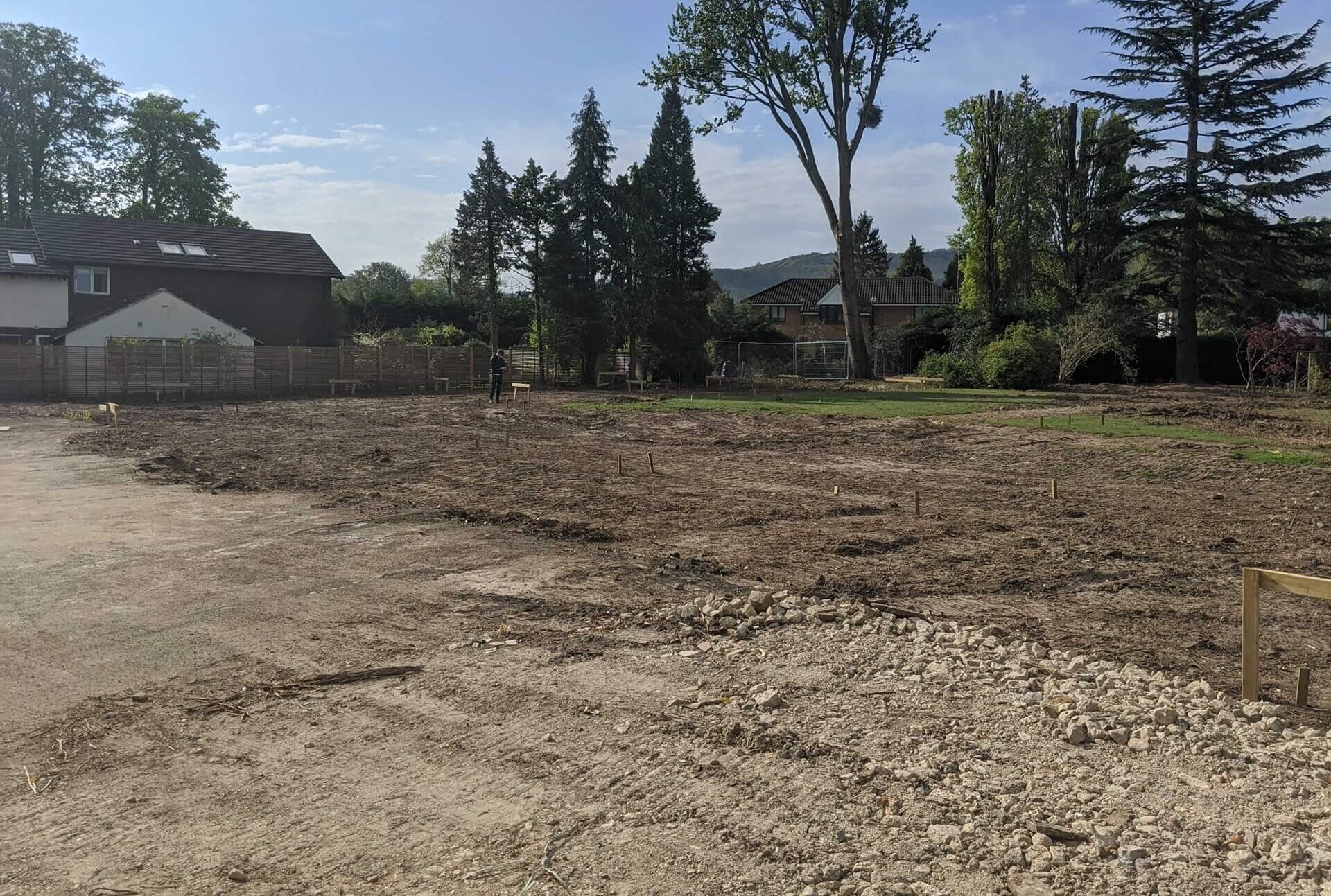 Bare garden site with gravel and rubble, a few mature trees at the edges, and houses beyond.