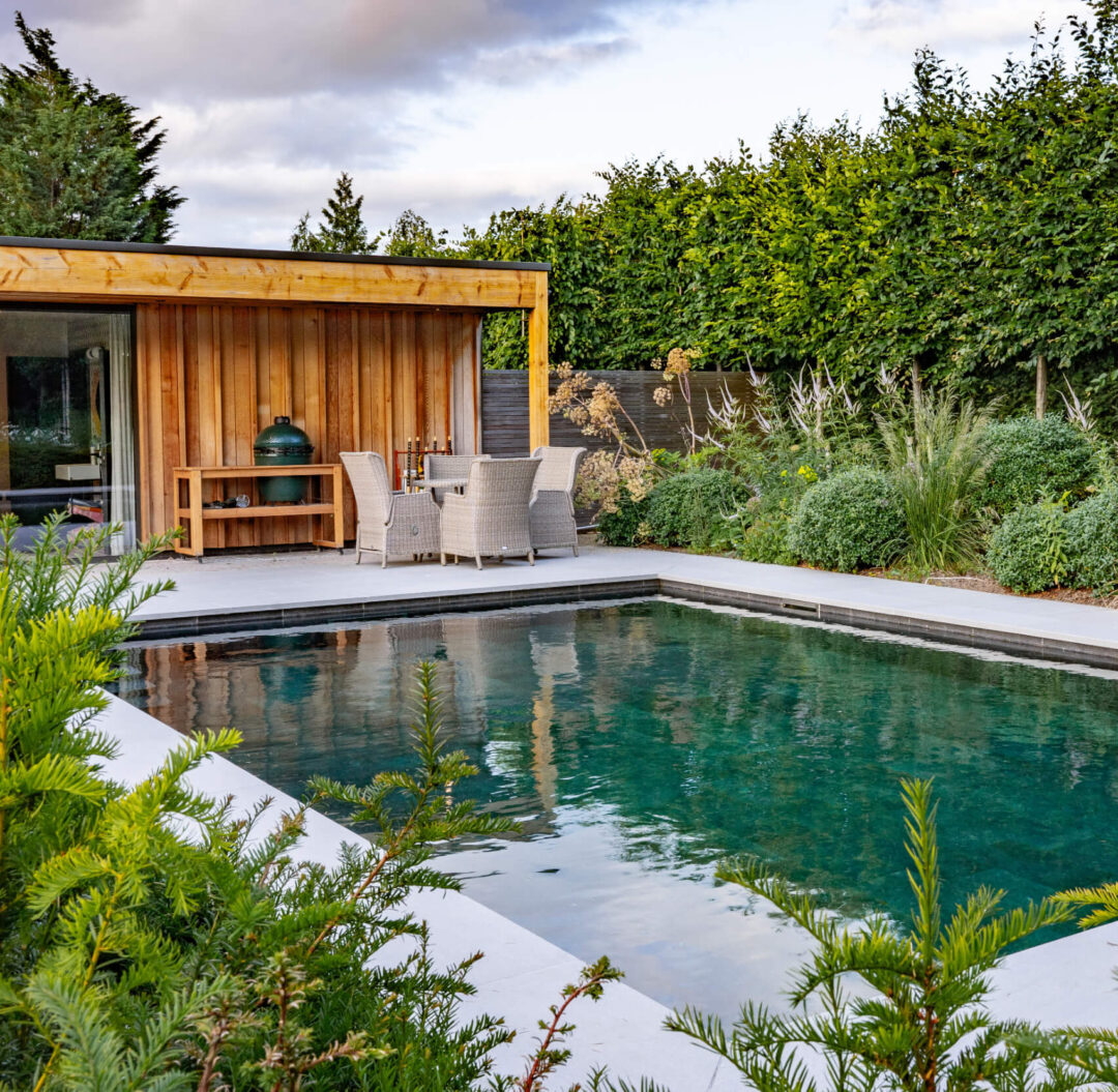 Shrubbery in the foreground with outdoor swimming pool behind, and warm-toned cedar pool house and hedging in the background.