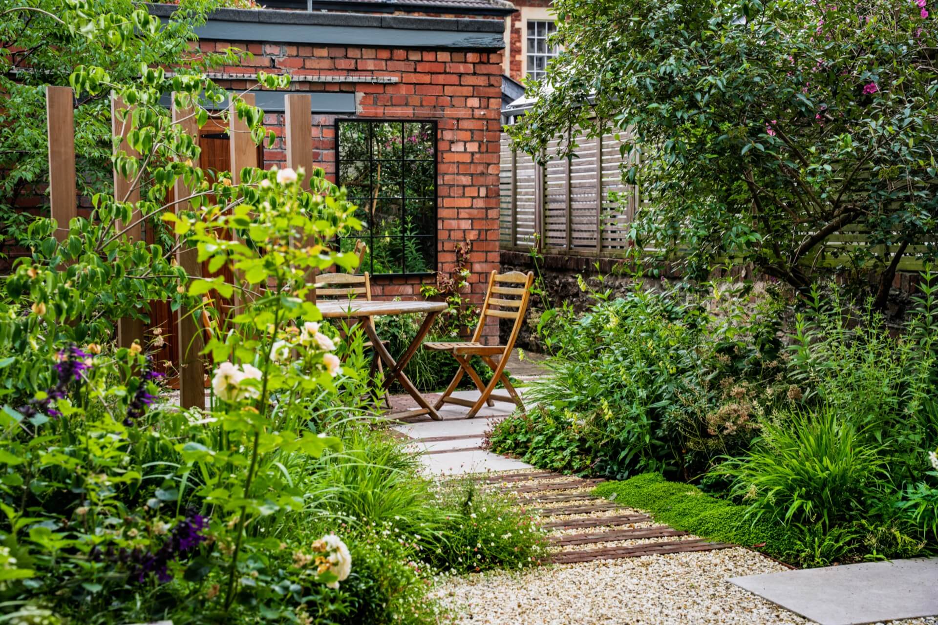 Cedar screening and uprights in a paved garden, with plenty of green foliage, and a dining area with wooden outdoor furniture towards the back. Red brick garage in the background with wooden door and window-style mirror.
