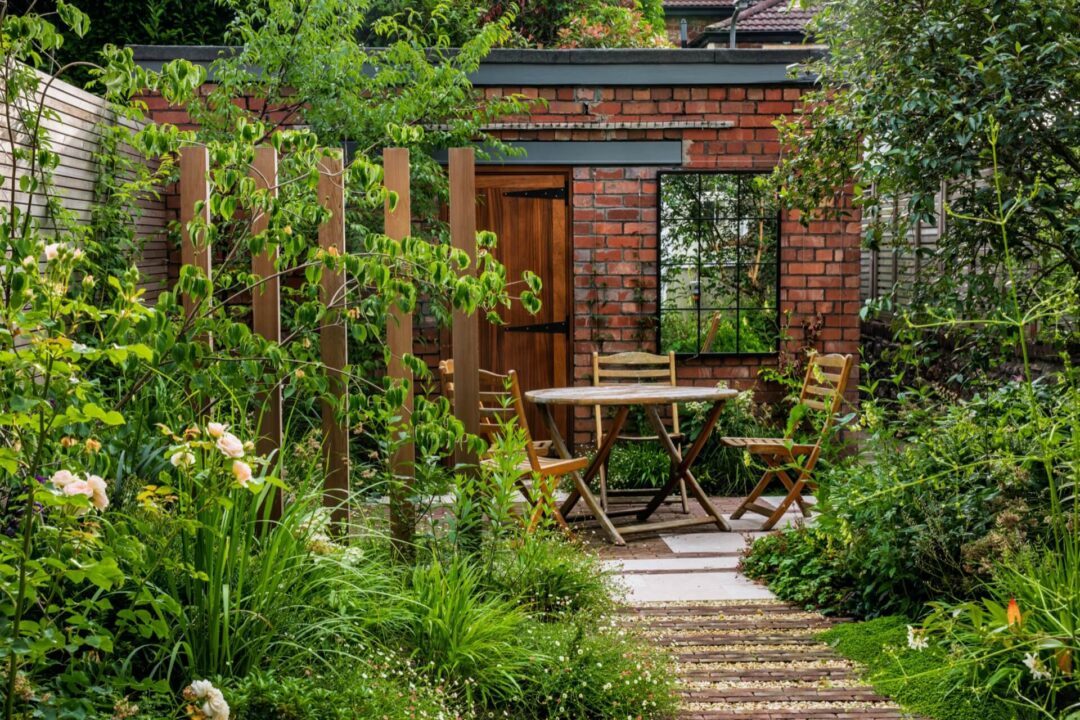 Cedar screening and uprights in a paved garden, with plenty of green foliage, and a dining area with wooden outdoor furniture towards the back. Red brick garage in the background with wooden door and window-style mirror.