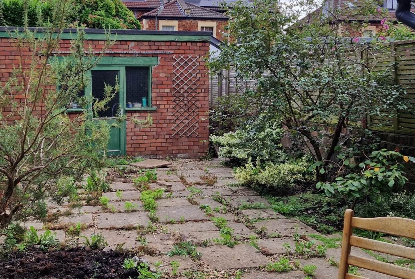 Paved garden with weeds and small trees/bushes, with red brick garage at the back with green wooden door and windows.